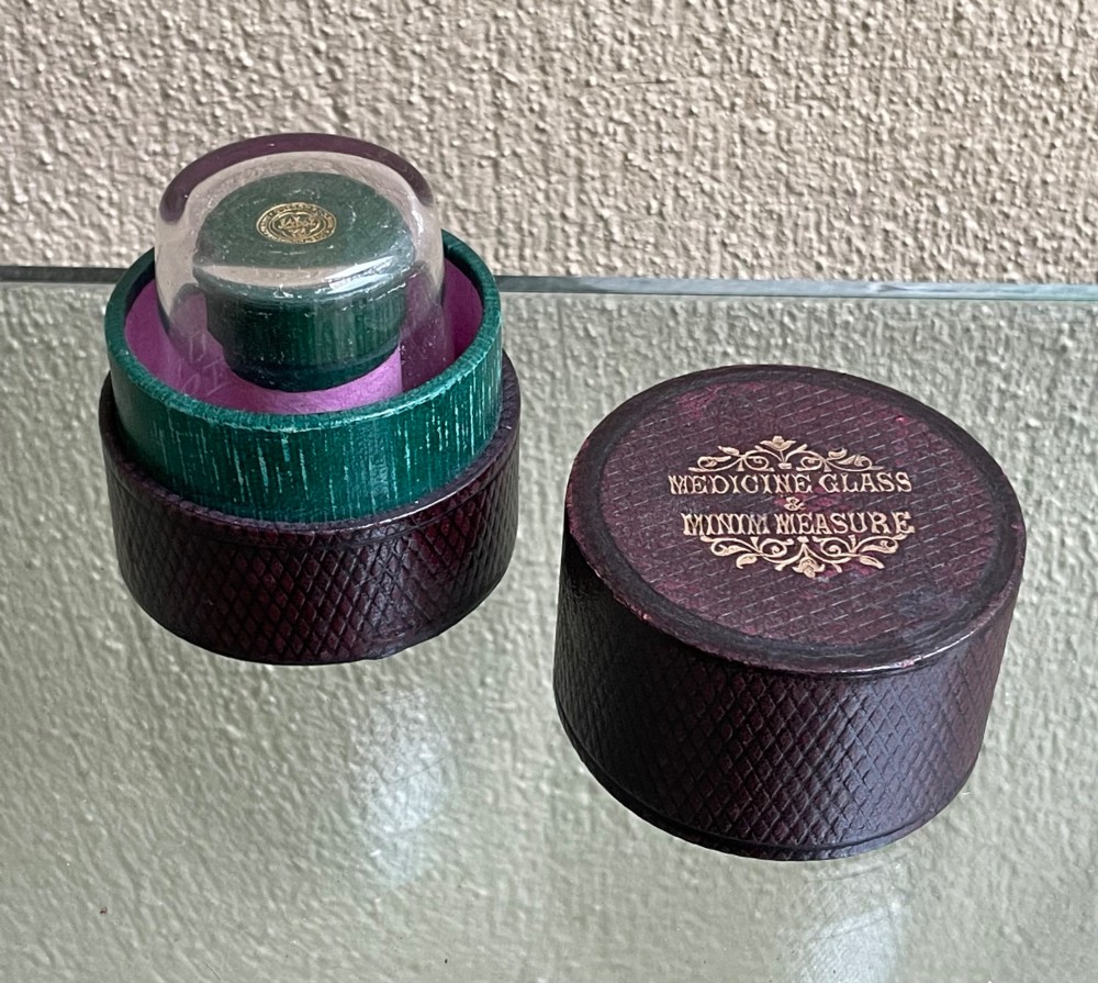 A 19th-century cylindrical burgundy textured leather case with gold embossed text reading Medicine Glass and Minim Measure alongside its fitted clear glass measuring cup.
