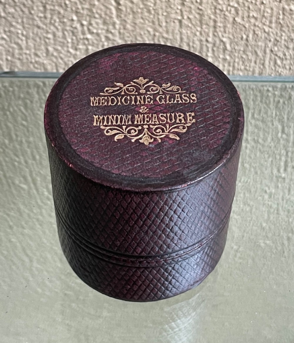 A 19th-century cylindrical burgundy textured leather case with gold embossed text reading Medicine Glass and Minim Measure alongside its fitted clear glass measuring cup. View II