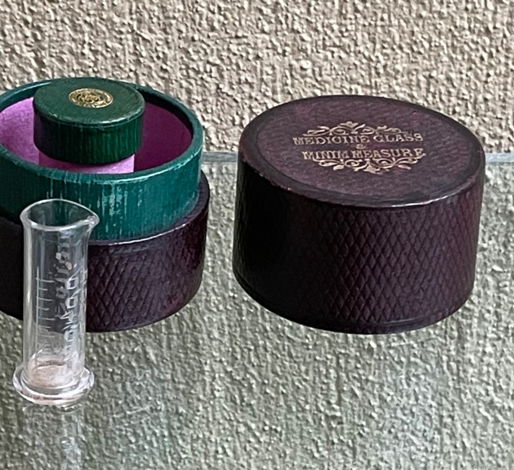 A 19th-century cylindrical burgundy textured leather case with gold embossed text reading Medicine Glass and Minim Measure alongside its fitted clear glass measuring cup. View VI