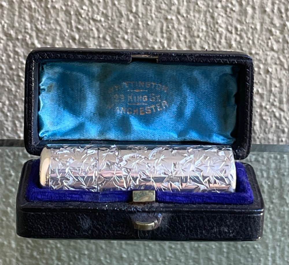 A cylindrical Victorian sterling silver scent bottle featuring chased foliage pattern work, resting inside an open leather presentation box lined with blue silk and velvet.