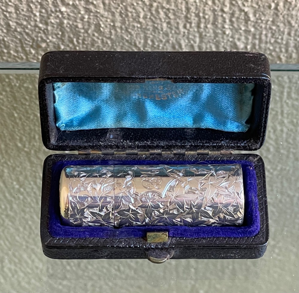 A cylindrical Victorian sterling silver scent bottle featuring chased foliage pattern work, resting inside an open leather presentation box lined with blue silk and velvet. View II