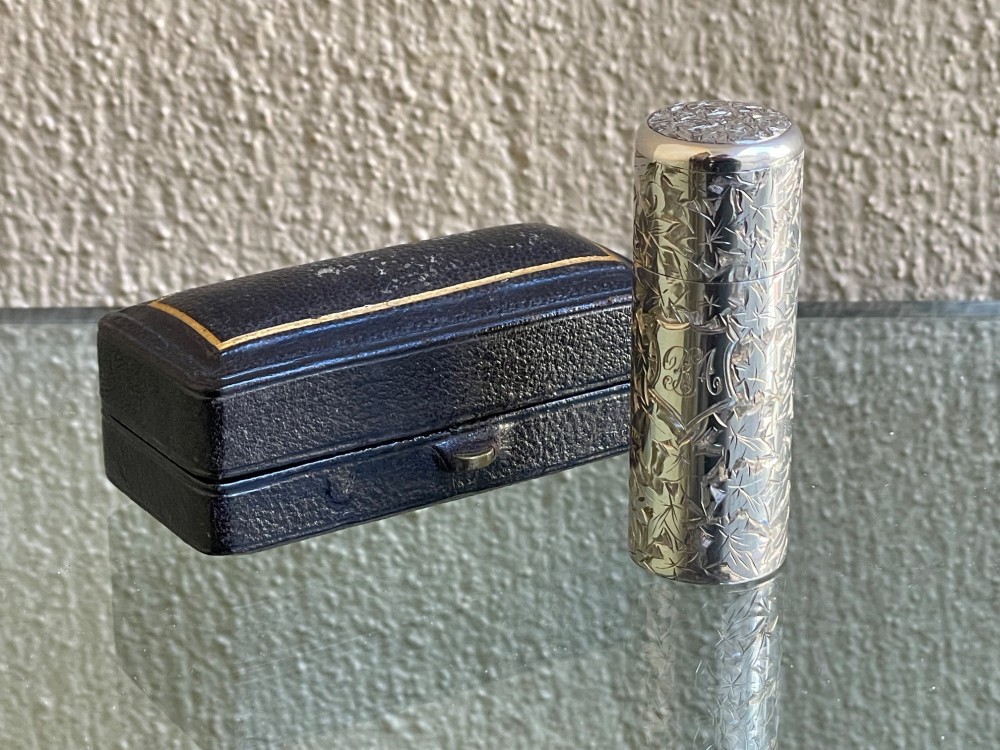 A cylindrical Victorian sterling silver scent bottle featuring chased foliage pattern work, resting inside an open leather presentation box lined with blue silk and velvet. View III