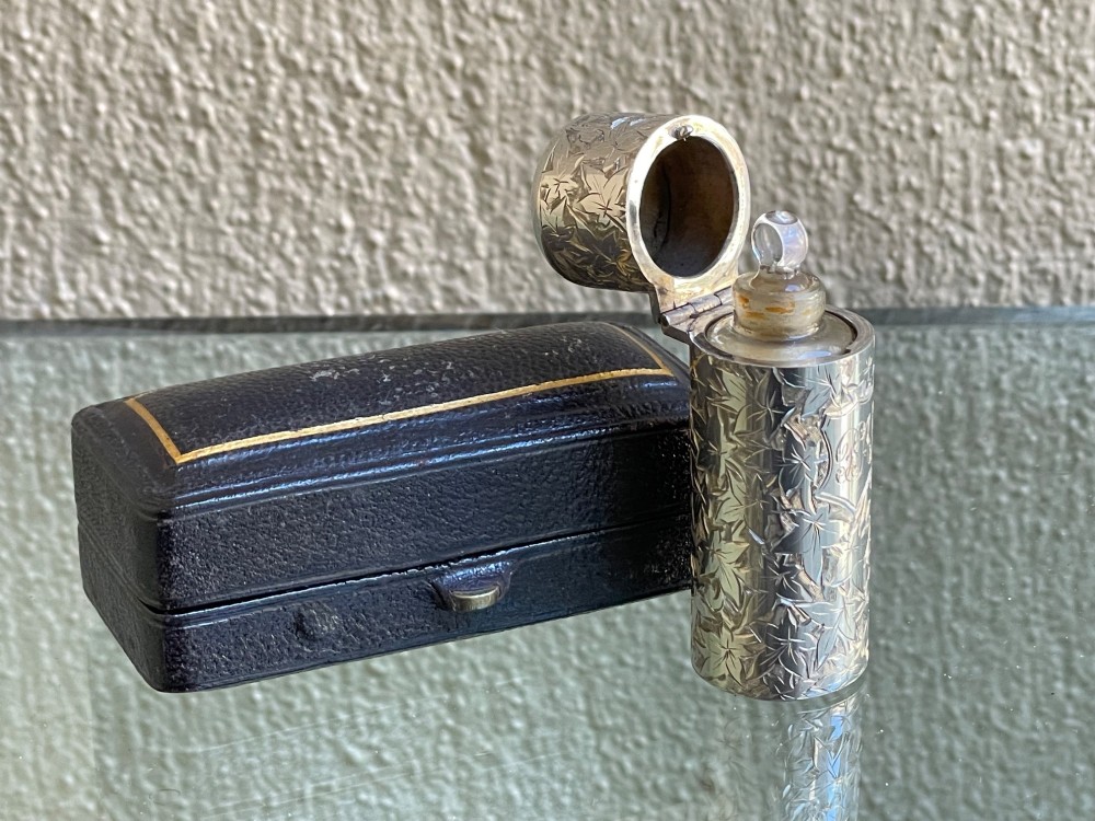 A cylindrical Victorian sterling silver scent bottle featuring chased foliage pattern work, resting inside an open leather presentation box lined with blue silk and velvet. View V