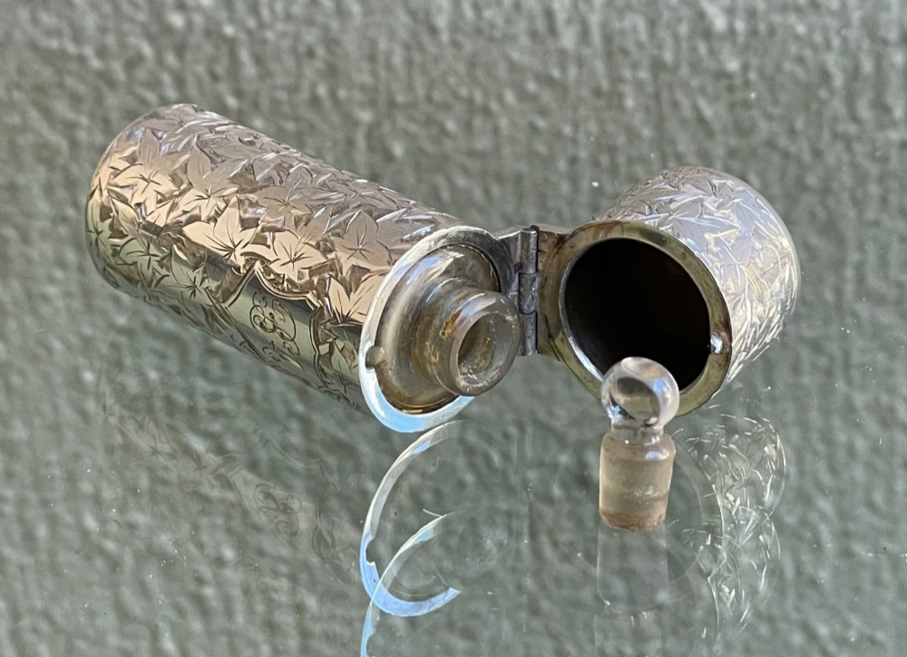 A cylindrical Victorian sterling silver scent bottle featuring chased foliage pattern work, resting inside an open leather presentation box lined with blue silk and velvet. View VI