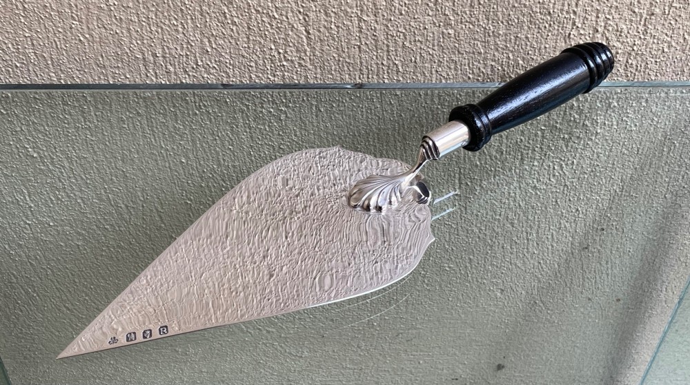 An antique Irish sterling silver presentation trowel with a textured engraved blade and turned black wooden handle, housed inside a black fitted presentation box with red lining. View II