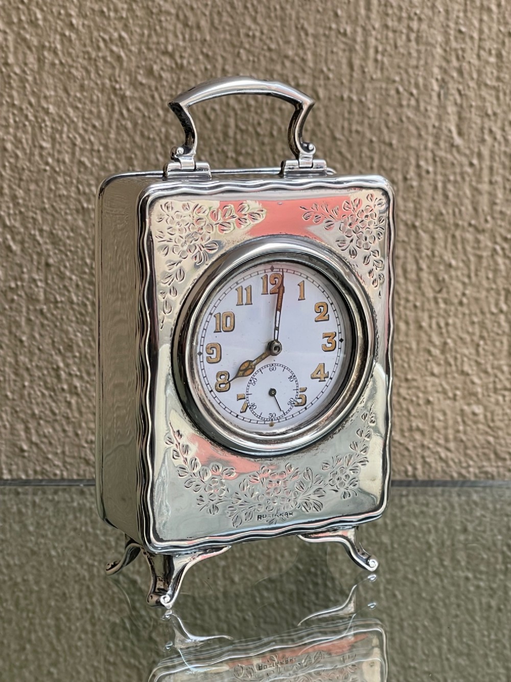 An Edwardian rectangular solid silver carriage clock featuring engraved floral foliage, a top carrying handle, cabriole feet, and a white enamel dial with Arabic numerals.