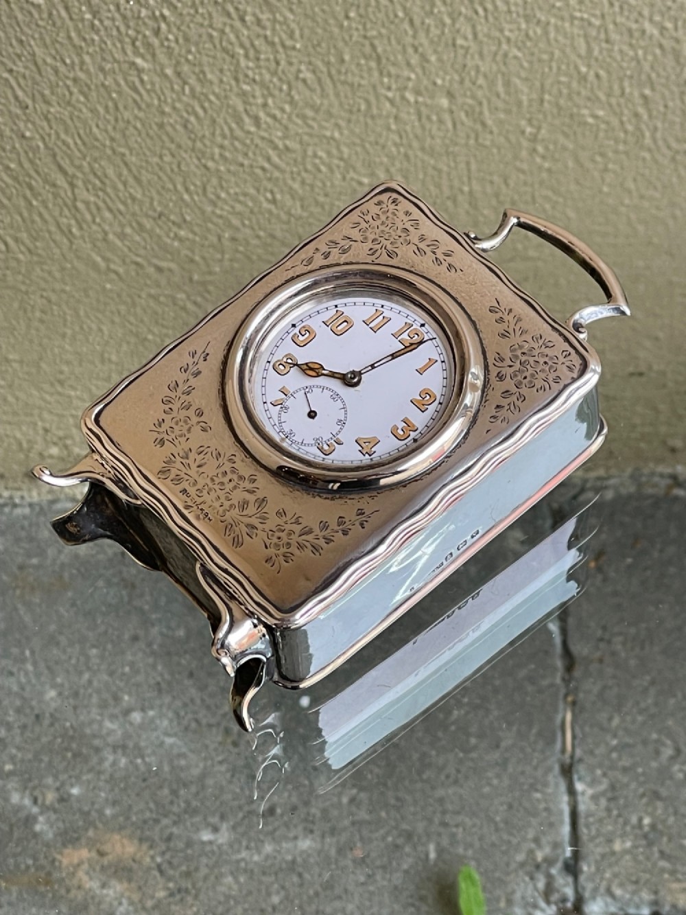 An Edwardian rectangular solid silver carriage clock featuring engraved floral foliage, a top carrying handle, cabriole feet, and a white enamel dial with Arabic numerals. View V