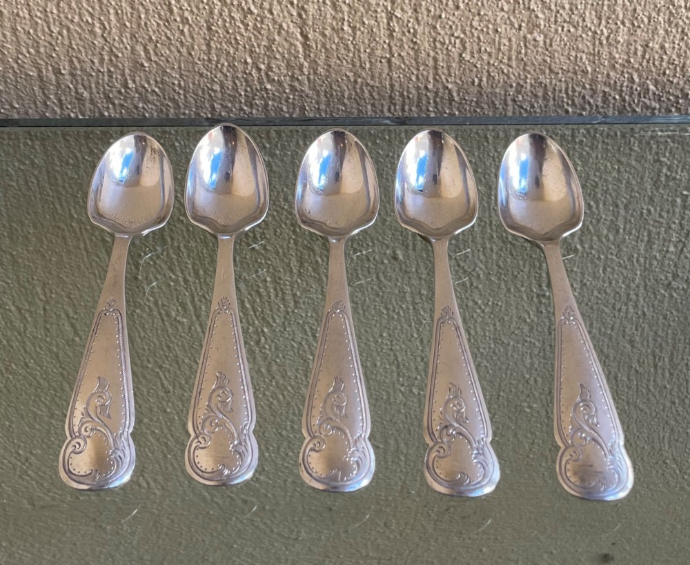 A set of five George V silver egg spoons featuring decorative engraved scrolled handles laid flat in a row.