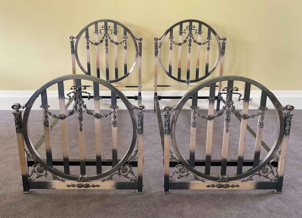 A matching pair of Art Nouveau nickel-plated single bed frames with circular slatted panels and cast ribbon swags.
