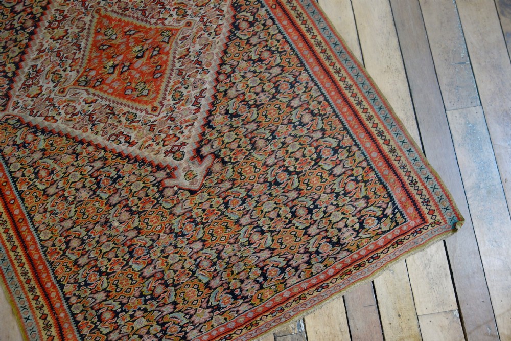 An antique hand-woven Senneh kilim rug featuring a prominent central diamond medallion surrounded by intricate repeating patterns in terracotta red, dark navy, and beige tones. View IV