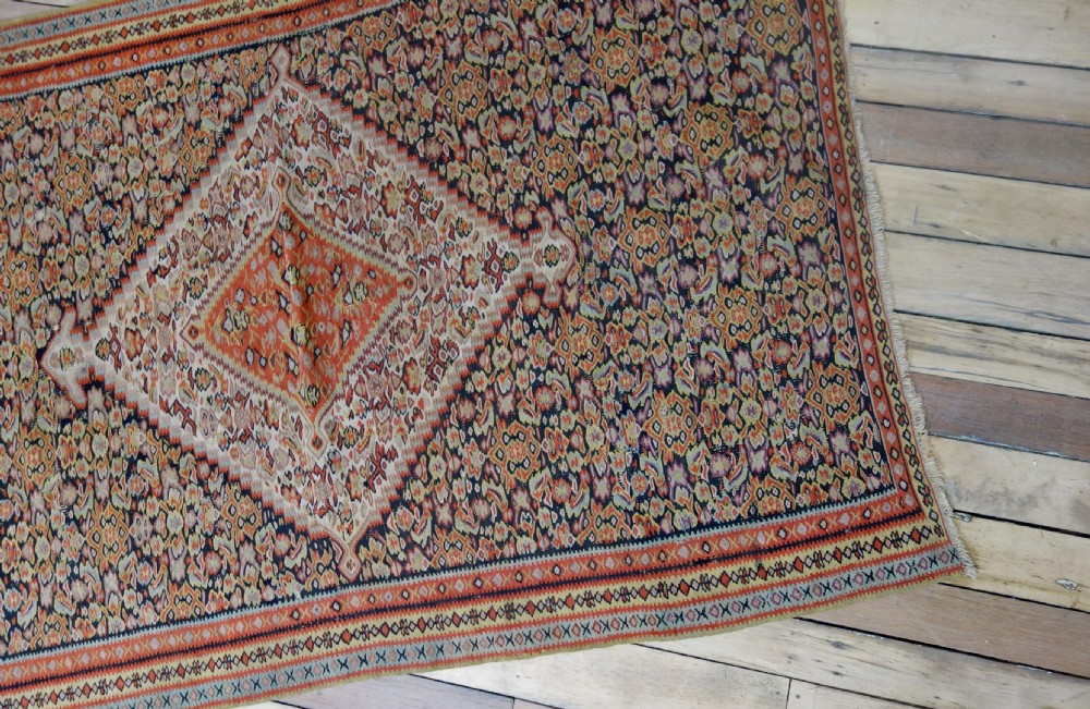An antique hand-woven Senneh kilim rug featuring a prominent central diamond medallion surrounded by intricate repeating patterns in terracotta red, dark navy, and beige tones. View V