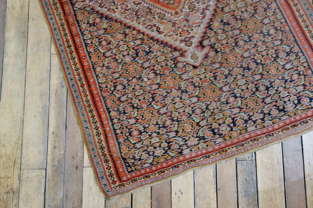 An antique hand-woven Senneh kilim rug featuring a prominent central diamond medallion surrounded by intricate repeating patterns in terracotta red, dark navy, and beige tones. View VI