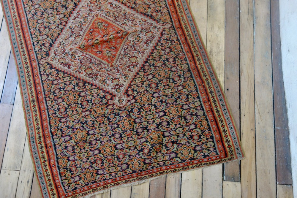 An antique hand-woven Senneh kilim rug featuring a prominent central diamond medallion surrounded by intricate repeating patterns in terracotta red, dark navy, and beige tones. View VII