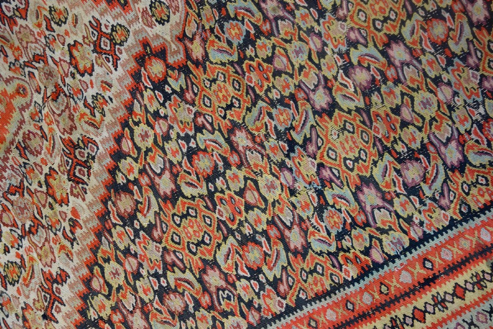 An antique hand-woven Senneh kilim rug featuring a prominent central diamond medallion surrounded by intricate repeating patterns in terracotta red, dark navy, and beige tones. View VIII