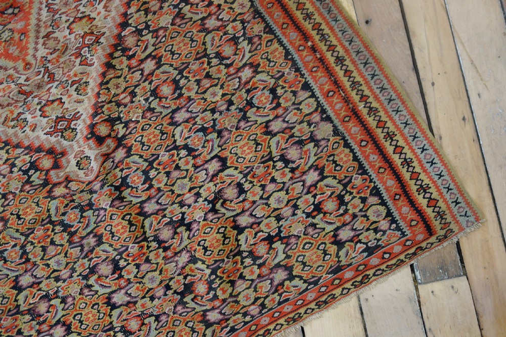 An antique hand-woven Senneh kilim rug featuring a prominent central diamond medallion surrounded by intricate repeating patterns in terracotta red, dark navy, and beige tones. View IX