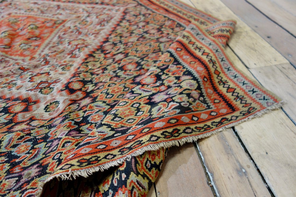 An antique hand-woven Senneh kilim rug featuring a prominent central diamond medallion surrounded by intricate repeating patterns in terracotta red, dark navy, and beige tones. View X