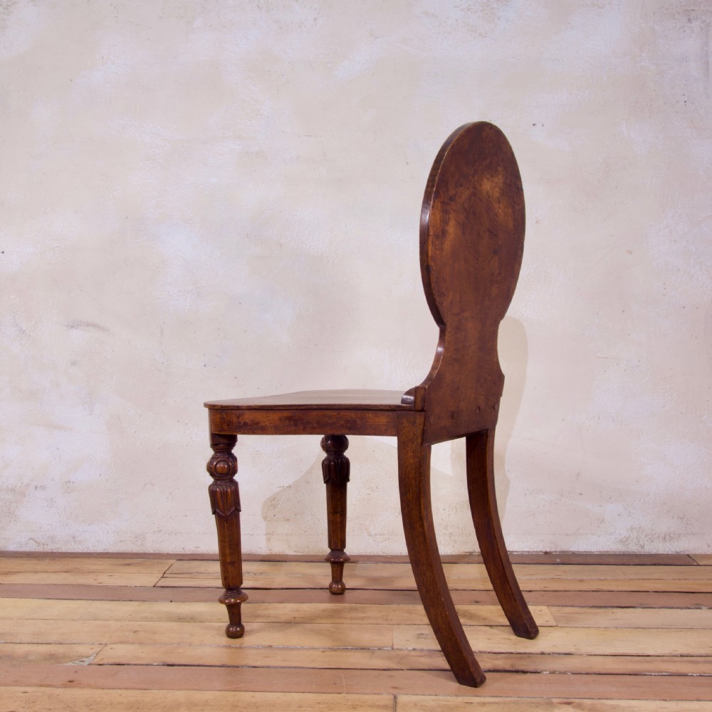 A 19th-century William IV hall chair made from elm and burr elm featuring a carved circular medallion back, solid wooden seat, turned front legs, and curved rear legs. View X