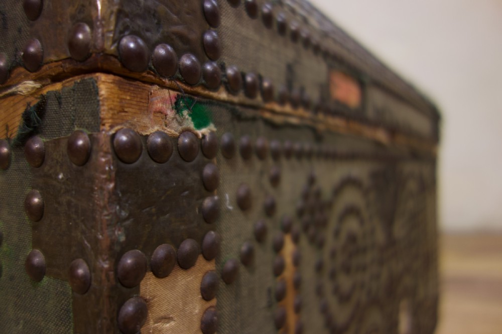 A small 19th-century French trunk featuring olive green fabric, brass stud patterning, iron side handles, and wooden bun feet. View V