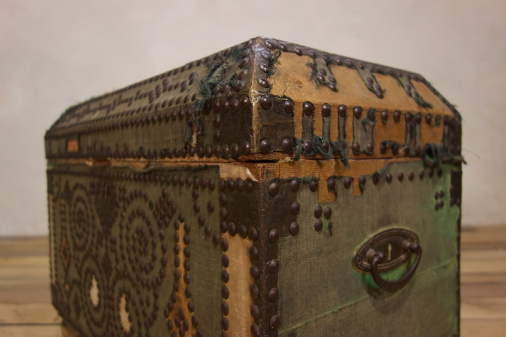 A small 19th-century French trunk featuring olive green fabric, brass stud patterning, iron side handles, and wooden bun feet. View X