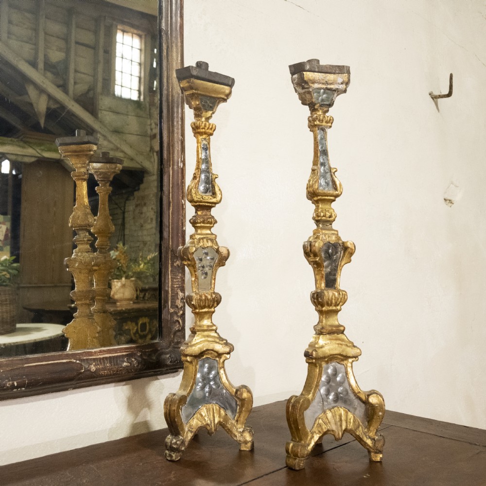 A pair of tall 18th-century Venetian giltwood altar candlesticks featuring carved details, inset mirrored glass panels, and triform bases.