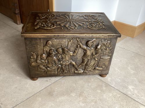 fig 1. An antique brass-clad wooden fireside log box featuring embossed relief scenes of figures in a tavern on the front and foliate scrollwork on the lid.