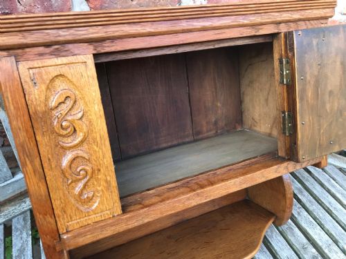fig 1. A Victorian carved oak wall cupboard featuring a central relief-carved door with bird motifs, flanked by scrolled side panels, a brass drop ring handle, and an open lower shelf.