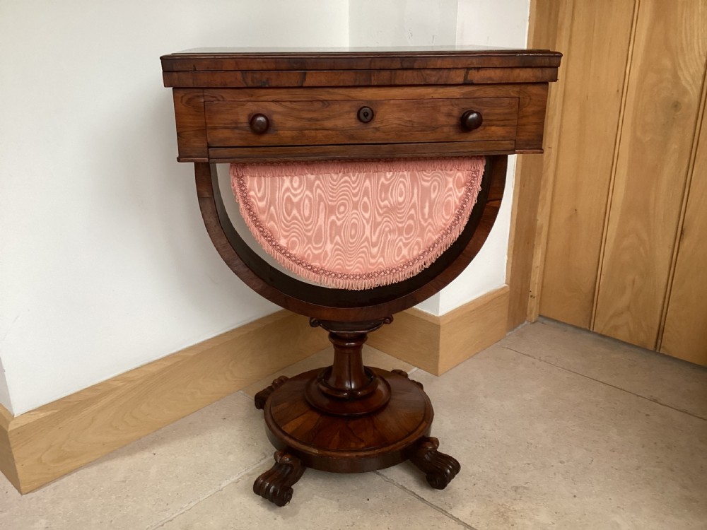 A 19th-century rosewood sewing table featuring a top drawer, a pink pleated silk work basket supported by a U-shaped frame, and a pedestal base with carved scroll feet.