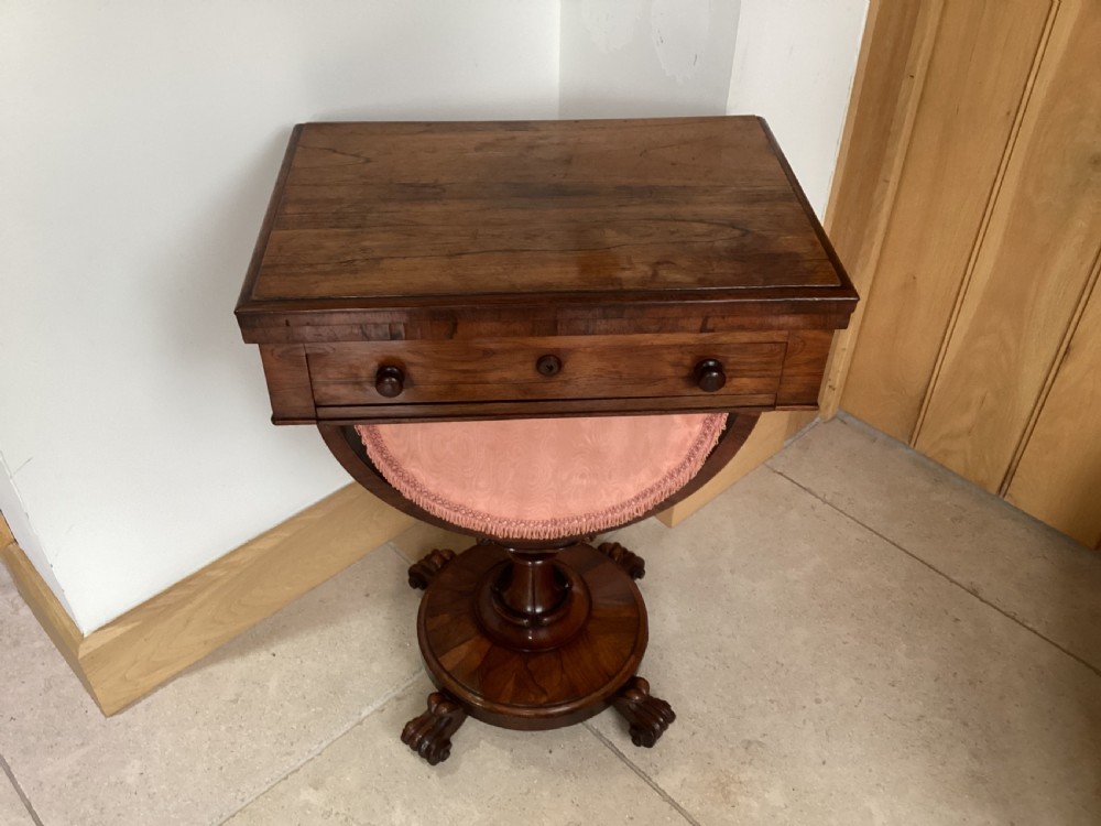 A 19th-century rosewood sewing table featuring a top drawer, a pink pleated silk work basket supported by a U-shaped frame, and a pedestal base with carved scroll feet. View X