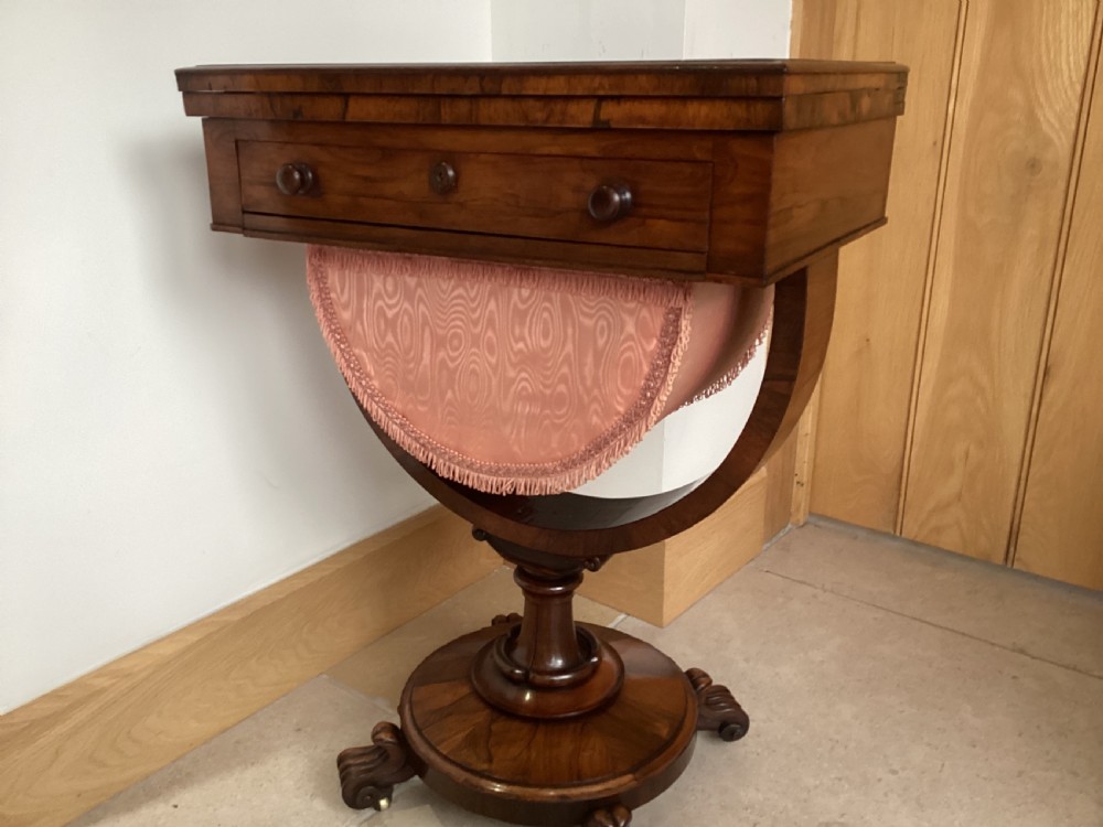 A 19th-century rosewood sewing table featuring a top drawer, a pink pleated silk work basket supported by a U-shaped frame, and a pedestal base with carved scroll feet. View XVI