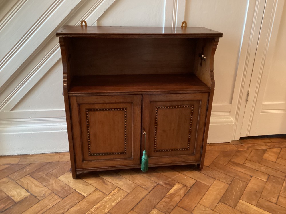 An Arts and Crafts mahogany wall cupboard featuring an open upper shelf, two inlaid lower doors, brass mounting brackets, and a key with a green tassel.