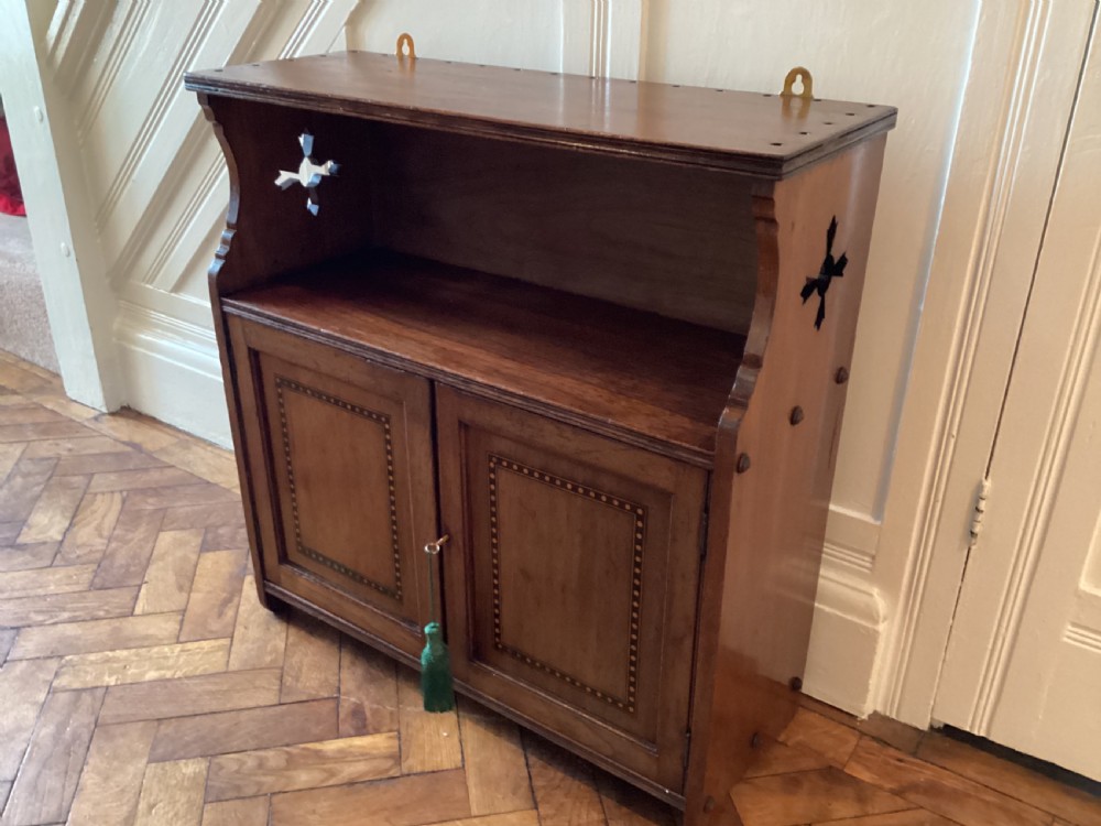 An Arts and Crafts mahogany wall cupboard featuring an open upper shelf, two inlaid lower doors, brass mounting brackets, and a key with a green tassel. View II
