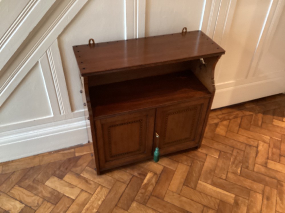 An Arts and Crafts mahogany wall cupboard featuring an open upper shelf, two inlaid lower doors, brass mounting brackets, and a key with a green tassel. View III