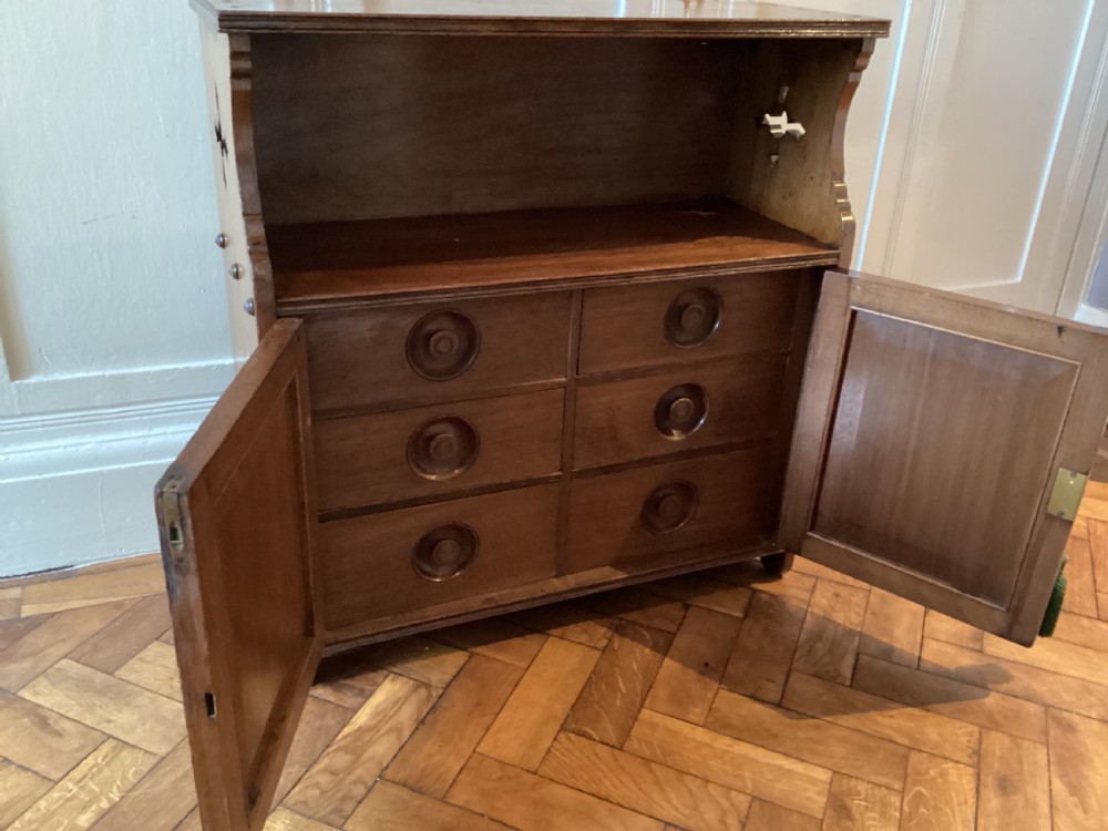 An Arts and Crafts mahogany wall cupboard featuring an open upper shelf, two inlaid lower doors, brass mounting brackets, and a key with a green tassel. View IV