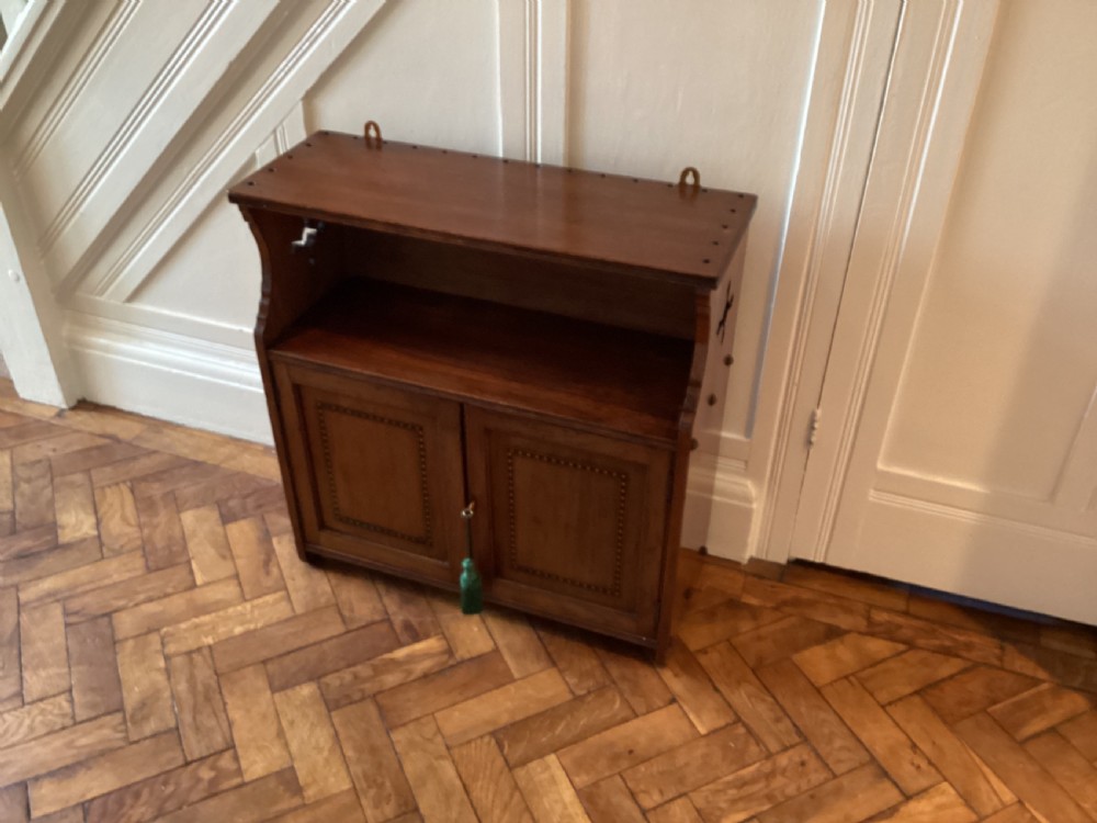 An Arts and Crafts mahogany wall cupboard featuring an open upper shelf, two inlaid lower doors, brass mounting brackets, and a key with a green tassel. View V