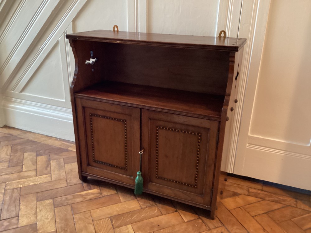 An Arts and Crafts mahogany wall cupboard featuring an open upper shelf, two inlaid lower doors, brass mounting brackets, and a key with a green tassel. View VI