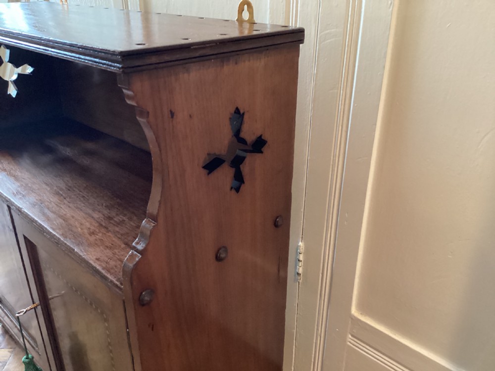 An Arts and Crafts mahogany wall cupboard featuring an open upper shelf, two inlaid lower doors, brass mounting brackets, and a key with a green tassel. View VII