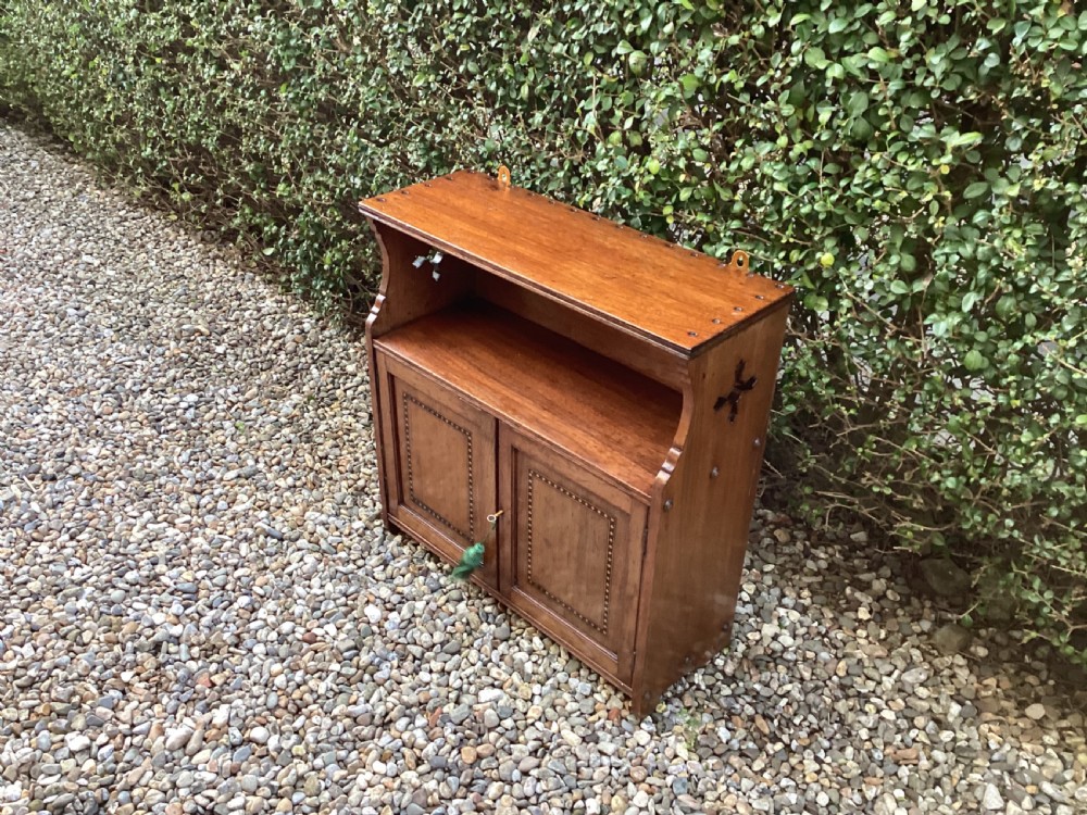 An Arts and Crafts mahogany wall cupboard featuring an open upper shelf, two inlaid lower doors, brass mounting brackets, and a key with a green tassel. View VIII