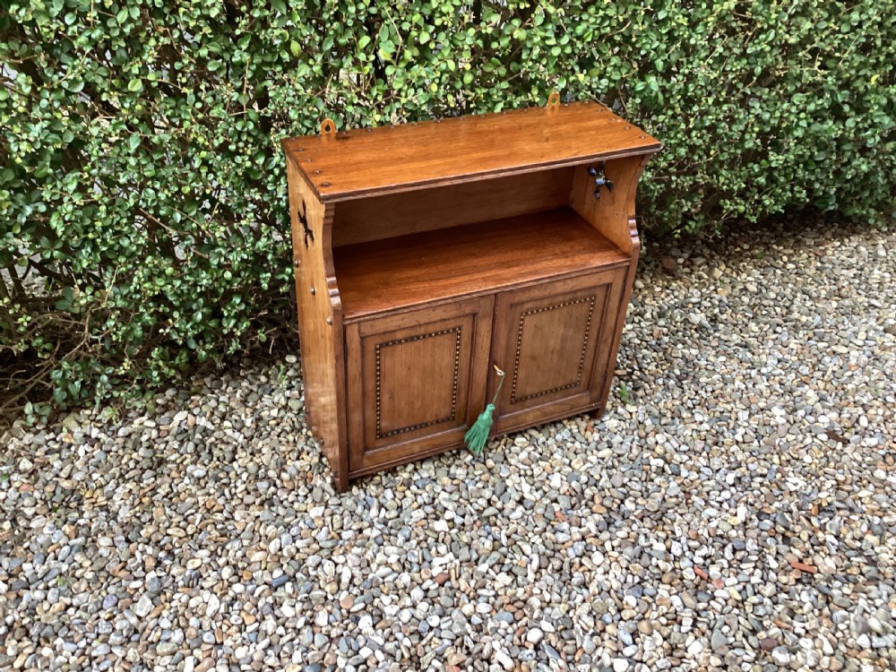 An Arts and Crafts mahogany wall cupboard featuring an open upper shelf, two inlaid lower doors, brass mounting brackets, and a key with a green tassel. View IX