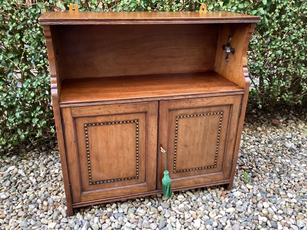 An Arts and Crafts mahogany wall cupboard featuring an open upper shelf, two inlaid lower doors, brass mounting brackets, and a key with a green tassel. View X