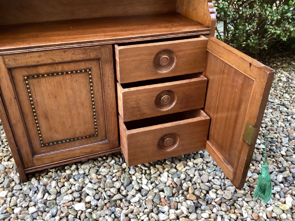 An Arts and Crafts mahogany wall cupboard featuring an open upper shelf, two inlaid lower doors, brass mounting brackets, and a key with a green tassel. View XI
