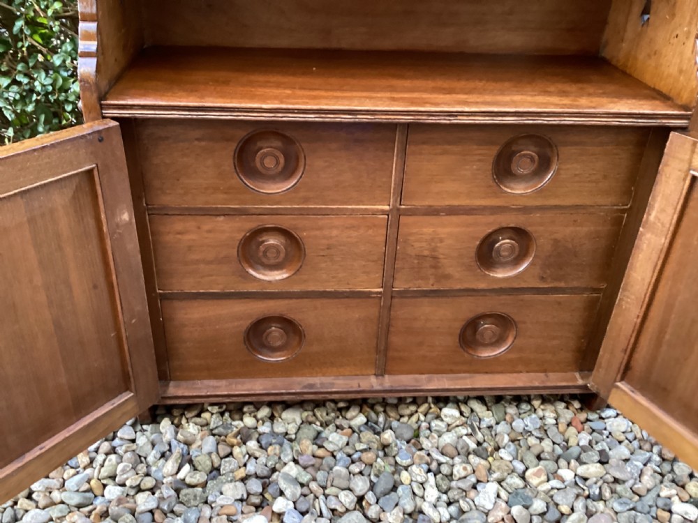 An Arts and Crafts mahogany wall cupboard featuring an open upper shelf, two inlaid lower doors, brass mounting brackets, and a key with a green tassel. View XII
