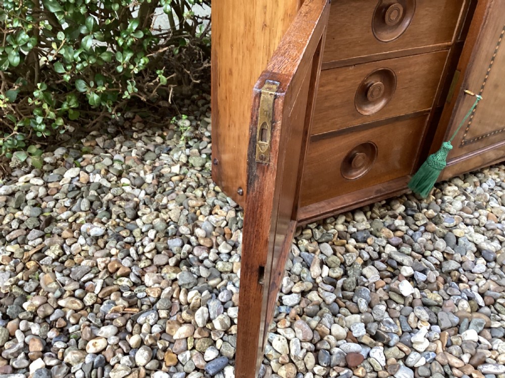 An Arts and Crafts mahogany wall cupboard featuring an open upper shelf, two inlaid lower doors, brass mounting brackets, and a key with a green tassel. View XIII