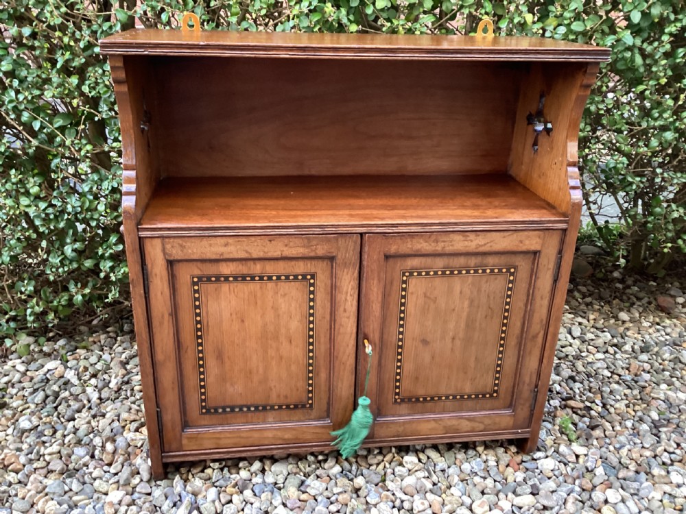 An Arts and Crafts mahogany wall cupboard featuring an open upper shelf, two inlaid lower doors, brass mounting brackets, and a key with a green tassel. View XVI