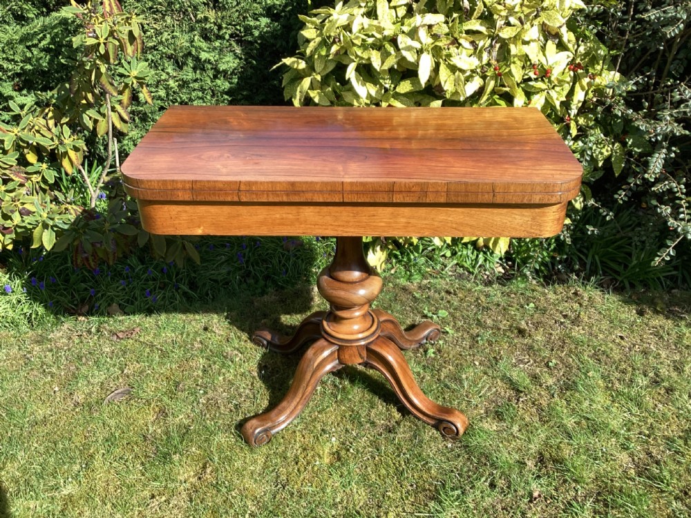 Victorian rosewood fold-over games table supported by a central turned pedestal column and four carved scroll feet.