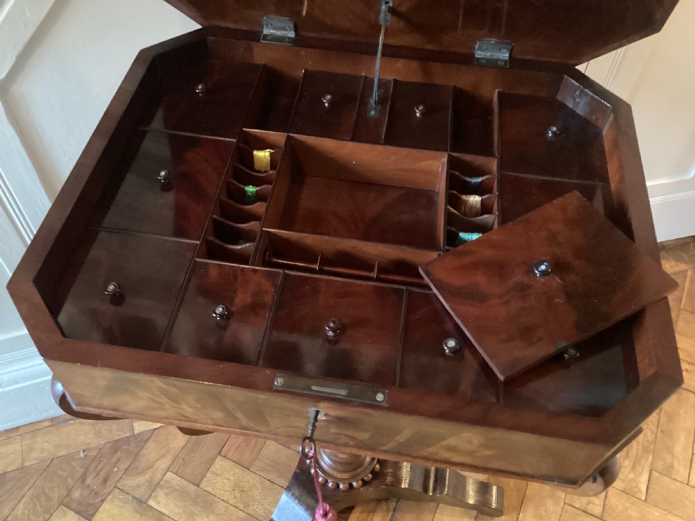 A Victorian flame mahogany sewing work table featuring a hinged top above a tapered work box, supported by curved S-scroll struts on a turned central pedestal with carved tripod legs. View VII