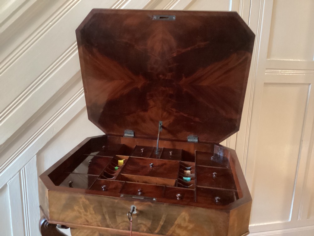 A Victorian flame mahogany sewing work table featuring a hinged top above a tapered work box, supported by curved S-scroll struts on a turned central pedestal with carved tripod legs. View XIX
