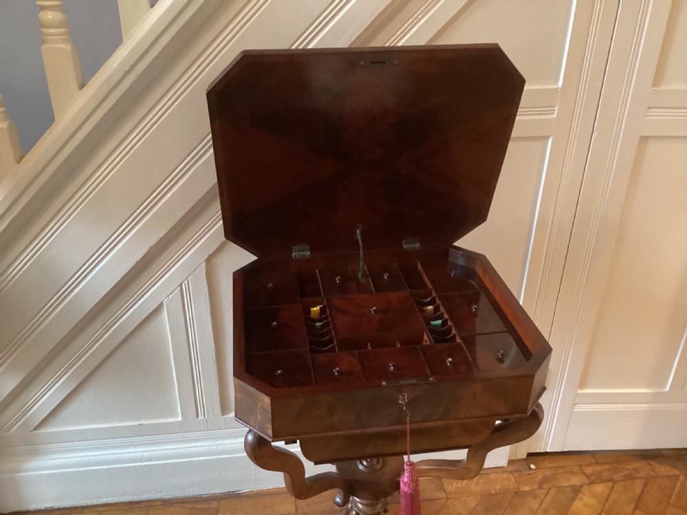 A Victorian flame mahogany sewing work table featuring a hinged top above a tapered work box, supported by curved S-scroll struts on a turned central pedestal with carved tripod legs. View XX