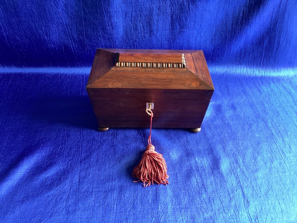 A Victorian rosewood tea caddy featuring a tapered sarcophagus lid with carved bead moulding, turned bun feet, and a central brass keyhole with a key and tassel. View X