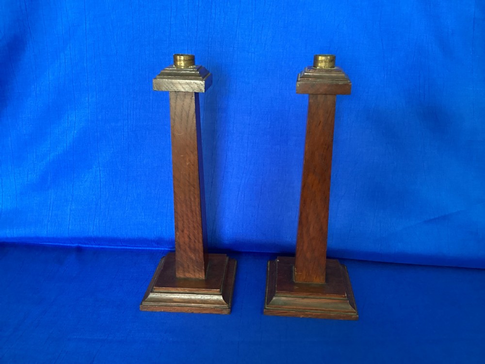 A pair of Edwardian Arts and Crafts tall oak candlesticks featuring square tapered shafts, stepped plinth bases, and brass nozzle inserts.