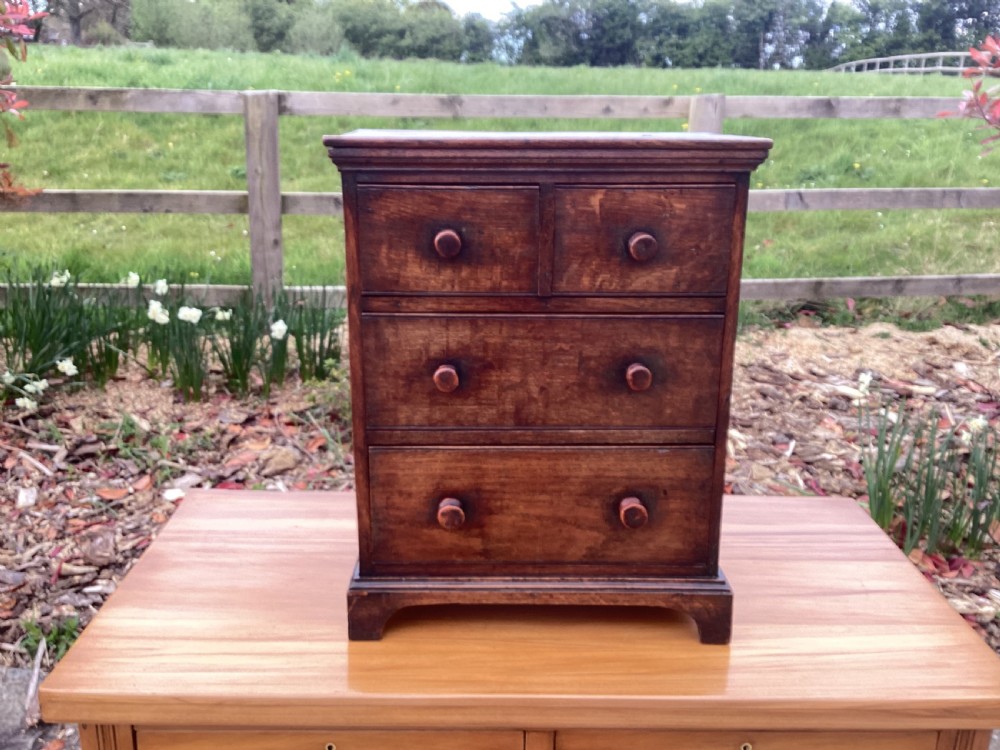 A small Georgian style miniature wooden chest of drawers featuring two short drawers over two long drawers with turned wooden knobs and bracket feet.